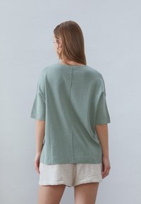 Top verde chiaro a maniche corte realizzato in tessuto morbido con una vestibilità comoda, caratterizzato da un dettaglio di cucitura sul retro. Abbinato a pantaloni corti bianchi con texture.