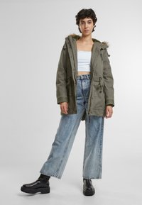 Oliegroene parka met een bontgevoerde capuchon, taillekoord en meerdere zakken. Gecombineerd met lichtblauwe wijde jeans en zwarte platformlaarzen.