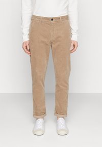 Pantalones de pana marrones con un corte recto, que cuentan con un cierre de botón y puños remangados. Zapatillas blancas con punta redonda completan el look.