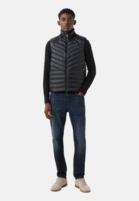 Czarna pikowana kamizelka bez rękawów z wysokim kołnierzem, dwie kieszenie na zamek, w połączeniu z granatową koszulą z długim rękawem i ciemnoniebieskimi dżinsami.
