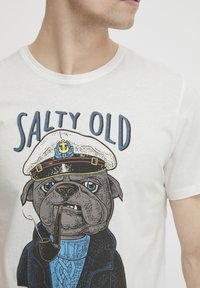 Hvid t-shirt med et grafisk motiv af en pug iført en kaptajnhat, blå sweater og sort frakke med teksten "SALTY OLD" ovenfor.