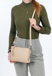 Borsa a tracolla in pelle beige chiaro con finitura testurizzata, dotata di chiusura con zip e tracolla regolabile. Indossata con un top olive a coste e pantaloni grigi.