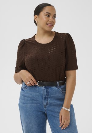 Femme portant un chemisier marron à manches courtes ajouré, rentrée dans un jean taille haute bleu avec une ceinture noire, souriante et regardant vers la droite.