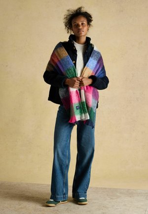 Young woman standing wearing a black jacket, white sweater, blue jeans, green shoes, and a colorful plaid scarf draped over her shoulders.