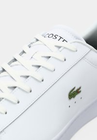 Bijele kožne tenisice s izraženim vezanjem, zelenim Lacoste krokodil logotipom i minimalističkim dizajnom. Imaju suptilne perforacije blizu prstiju.