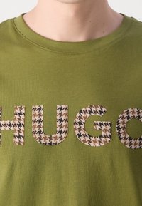 Zbliżenie na osobę w oliwkowej koszuli z dużymi literami "HUGO" w brązowym i czarnym wzorze w zęby psa na klatce piersiowej.