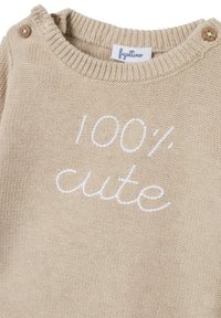 Pull en tricot beige avec un col rond, des accents de boutons en bois sur l'épaule et un texte brodé indiquant "100 % mignon" en blanc.