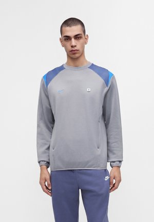 Jeune homme aux cheveux courts portant un haut gris à manches longues avec des accents bleus aux épaules et un pantalon de survêtement Nike bleu, debout devant un fond uni.