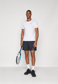 White athletic t-shirt with short sleeves and a black logo; paired with black shorts. The model holds a tennis racket and wears black sneakers.