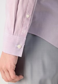 Gestreiftes Hemd in Hellpink und Weiß mit knöpfbarem Umschlag, ausgestattet mit einem kleinen Logo und einer glatten Textur. Getragen mit grauen Hosen.