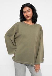 Ελαιόλαδο πράσινο oversized φούτερ με μακριά, φαρδιά μανίκια, στρογγυλή λαιμόκοψη και λευκές ραφές κατά μήκος του στριφώματος και των μανικιών.