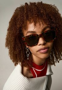 Tortoiseshell zonnebril met een vierkant montuur en donkere lenzen, gecombineerd met een wit geribbeld jasje en een rode top met witte afwerking.