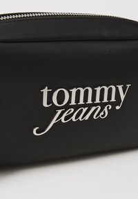 Juodas dirbtinės odos krepšys su sidabru „tommy jeans“ logo, užtrauktuku ir lygiu paviršiumi. Kompaktiškos stačiakampio formos su subtiliomis detalėmis.