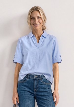 Femme blonde souriante, portant une chemise bleu clair à manches courtes avec col et un jean bleu, debout devant un mur clair uni.