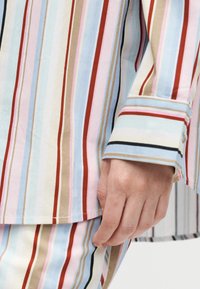 Χέρι ακουμπά κοντά στο ισχίο, φοράει ρούχο με κάθετες ρίγες σε κόκκινο, μπλε, μπεζ, ροζ και μαύρο σε ανοιχτό φόντο.