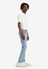 Hvit kortermet t-skjorte, lyseblå rett ben jeans og hvite joggesko med brune detaljer. Modellen står sidelengs mot en hvit bakgrunn.