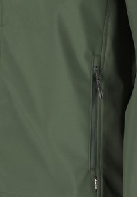 Vihreän takin osa, jossa pystysuuntainen vetoketjullinen tasku, jossa on kaksi vetimetappia ja pieni kangasmerkki lähellä helmaa.