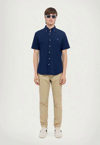 Άνδρας με ναυτικό μπλε κοντομάνικο πουκάμισο, μπεζ παντελόνι, λευκά αθλητικά και γυαλιά ηλίου, που στέκεται μπροστά σε λευκό φόντο.