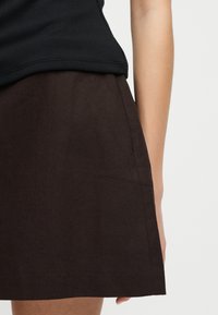 Γυναίκα με σκούρο καφέ φούστα και μαύρο ριμπ τοπ, εμφανιζόμενη από τη μέση έως το μηρό σε απλό φωτεινό φόντο.