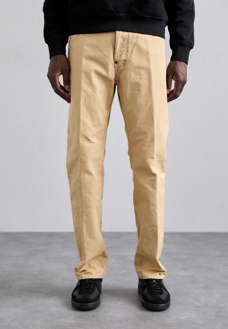 Pantalon en coton beige avec une coupe droite, doté d'une fermeture à bouton et de poches latérales, associé à des baskets noires.