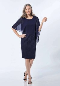 Navy Kleid mit einer durchsichtigen, geschichteten Cape. Strukturiertes, plissiertes Gewebe in einer figurbetonten Silhouette. Offenporige schwarze Sandalen runden den Look ab.