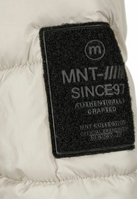 Bežs pūkains jaka audums ar melnu, teksturētu birku, kurā ir iembossēts teksts: "MNT SINCE 97", "AUTHENTICALLY CRAFTED" un logotips.