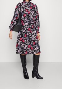 Černé květinové šaty s červenými, fialovými a bílými vzory; dlouhé rukávy, vysoký límec; doplněné černými koženými botami po kolena a černou taškou.