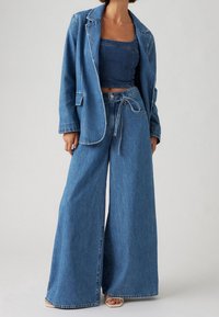 Denimdräkt med skräddarsydd jacka och vida byxor. Tyget är medelblått med kontrasterande sömmar och bältesdetalj på byxorna.