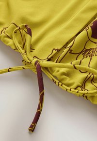 Żółty materiał z kwiatowym wzorem, z marszczonym designem i troczkami w kolorze bordowym. Gładka tekstura i miękkie wykończenie.