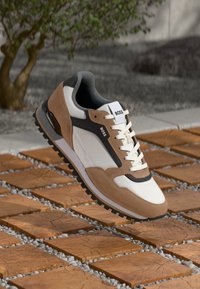 Beige en witte sneaker met een textuur bovenkant, zwarte accenten en een rubberen zool. Voorzien van veters en een merklabel op de tong.