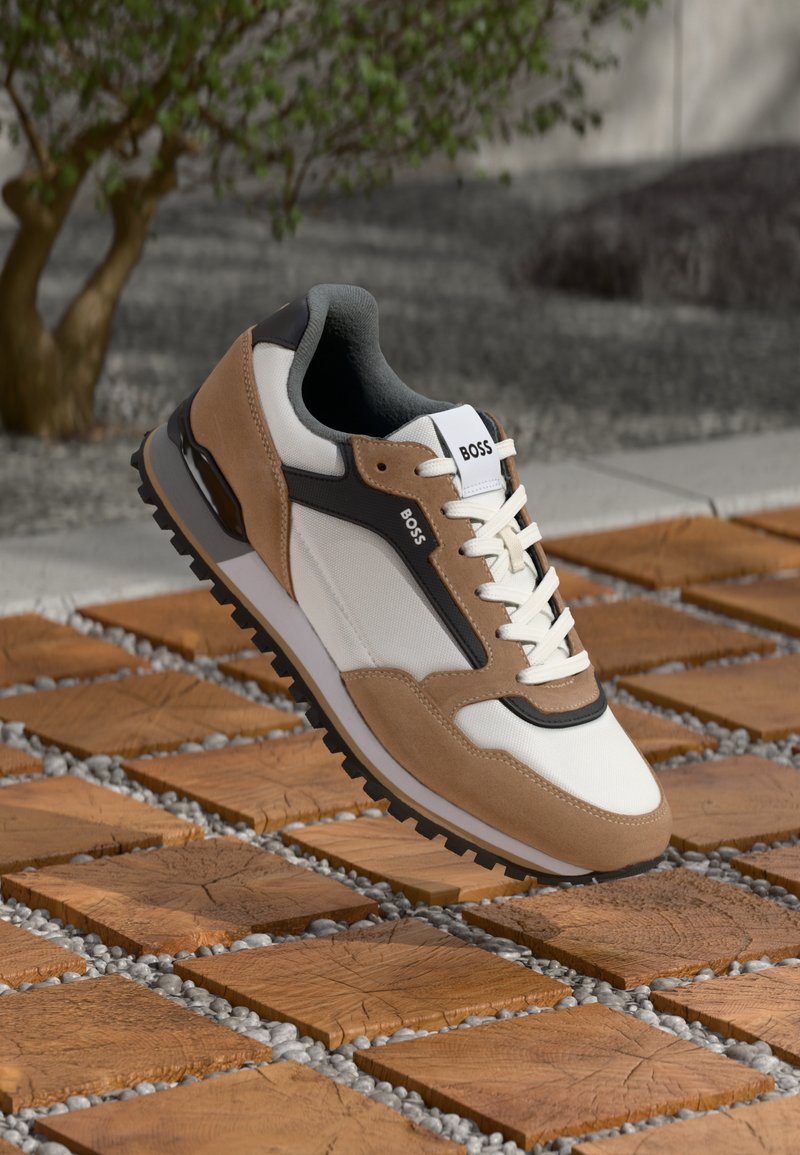 Beige és fehér sneaker, texturált felsőrésszel, fekete kiegészítőkkel és gumitalppal. Fűzőkkel rendelkezik, valamint márkajelzés található a nyelvén.