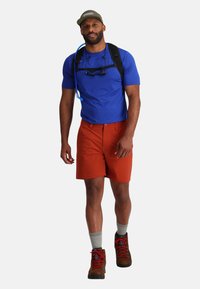 Maglietta sportiva blu, pantaloni corti arancioni, calze grigie e scarponi da trekking marroni con lacci rossi. Indossa un cappellino verde e porta uno zaino.