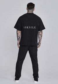 Czarny t-shirt z białym logo "SIKSILK" na plecach, w połączeniu z czarnymi spodniami. Materiał wydaje się miękki; krój jest luźny z krótkimi rękawami.