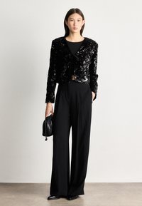 Μαύρο κομμένο μπλέιζερ με παγιέτες, κλείσιμο με κορδόνι μπροστά, φορεμένο πάνω από ένα μαύρο τοπ και συνδυασμένο με μαύρα παντελόνια ευρείας γραμμής. Περιλαμβάνει μια μικρή μαύρη τσάντα.