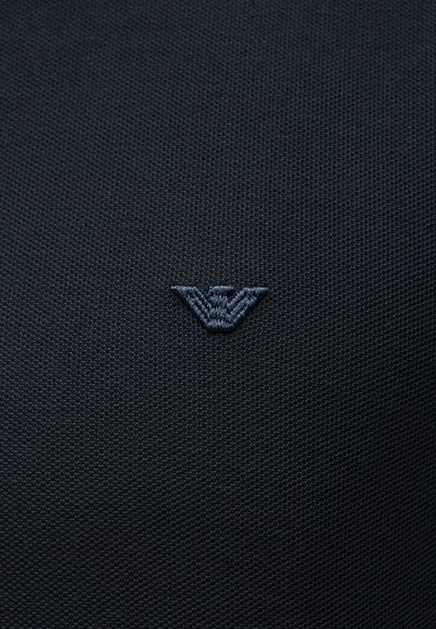 Σκούρο υφασμάτινο ύφασμα με μικρό, κεντρικά τοποθετημένο κεντημένο στυλιζαρισμένο λογότυπο αετού σε ελαφρώς πιο ανοιχτή απόχρωση.