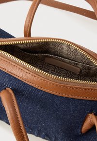 Riflová taška s hnědými koženými detaily, zlatým zipem a vzorovanou vnitřní podšívkou. Má pevné popruhy a kompaktní, strukturovaný design.