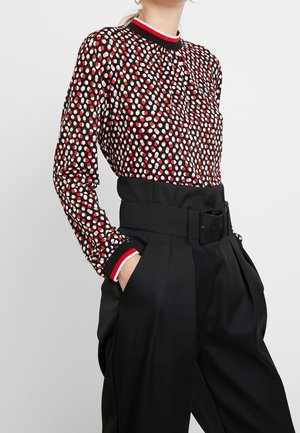 Femme portant un chemisier noir à manches longues à pois blancs et rouges et un pantalon noir taille haute avec une ceinture large, les mains dans les poches.