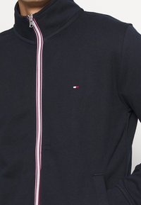 Granatowa kurtka zapinana na zamek z wysokim kołnierzem, ozdobiona czerwono-białymi paskami biegnącymi wzdłuż zamka oraz małym logo na piersi. Miękka tekstura materiału.