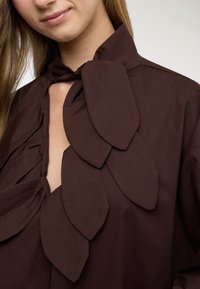Blusa marrone con collo alto e grandi decorazioni a forma di foglia sulla parte anteriore. Il tessuto sembra liscio con una lucentezza sottile.