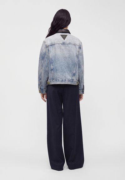 Femme aux longs cheveux foncés portant une veste en jean bleu clair et un pantalon large noir, debout face à un fond uni gris clair.