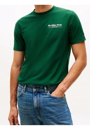 Mężczyzna w zielonej koszulce z krótkim rękawem z napisem "The Hilfiger Herald" i niebieskich dżinsach, z jedną ręką w kieszeni na białym tle.