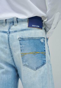 Jeans de mezclilla azul claro con un bolsillo desgastado y costuras amarillas, además de una etiqueta trasera que dice "SALSA JEANS". Diseño clásico de cinco bolsillos.