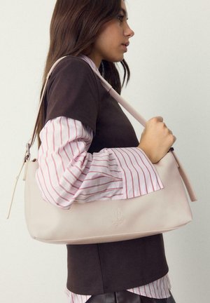 Mladá žena v vrstveném hnědém topu a proužkované košili drží světlé béžové kabelce na rameno s logem na jednobarevném pozadí.