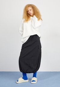 Sweatshirt bianco oversize, gonna cargo nera con bordo elastico, calzini blu a costine e sandali beige scivolabili su un pavimento blu chiaro.