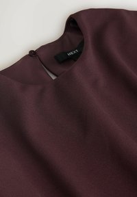 Burgundská blůza s hladkým povrchem, zaobleným výstřihem a zapínáním na knoflík na zadní straně; obsahuje štítek s názvem značky "NEXT."