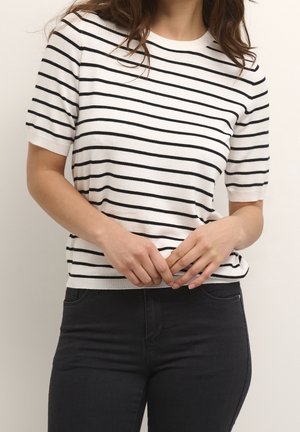Frau trägt ein weißes kurzärmeliges Hemd mit schwarzen horizontalen Streifen und schwarze Hose, Hände nahe der Taille verschränkt.