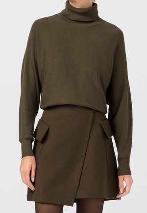 Femme portant un pull à col roulé marron et une jupe marron assortie avec des poches rabatées à l’avant, debout devant un fond uni.