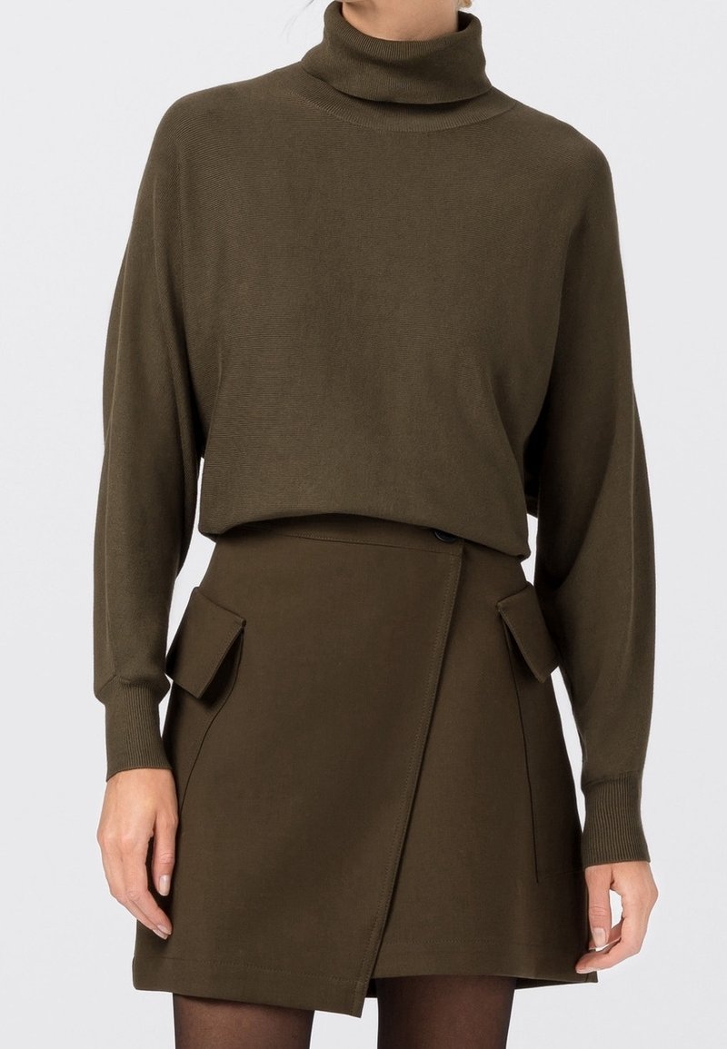 Femme portant un pull à col roulé marron et une jupe marron assortie avec des poches rabatées à l’avant, debout devant un fond uni.