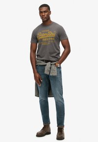 Graues T-Shirt mit gelbem Text, blaue Jeans und braune Schnürstiefel. Ein kariertes Hemd, das um die Taille gebunden ist, sorgt für Struktur und einen Mustermix.