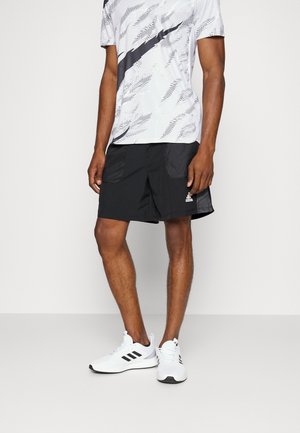 Mężczyzna w białej i czarnej wzorzystej koszulce Nike, czarnych sportowych szortach Adidas i białych butach sportowych Adidas, stojący na jednolitym tle.